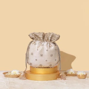 Può includere: Una borsa a sacchetto color crema con un intricato ricamo argentato a motivo floreale. La borsa presenta una parte superiore arricciata, cordini intrecciati argentati con nappe di perline e una finitura in tessuto testurizzato con dettagli metallici.