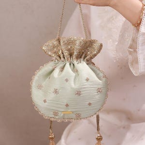May include: A light mint green drawstring pouch bag featuring intricate gold embroidery, small floral motifs, and sequin embellishments. The bag has a gathered top, a braided gold handle, and two dangling gold beaded tassels at the bottom base.