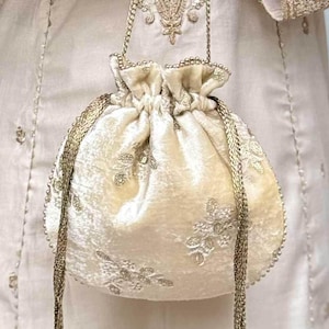 Può includere: Una borsa a sacchetto in velluto color crema con ricami floreali color oro, un manico a catena dorata e due lunghi cordini intrecciati dorati che terminano con nappe decorative di perline color oro scuro sul fondo della borsa.