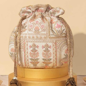 Peut inclure: Un sac à cordon blanc avec un motif floral et des broderies dorées. Le sac a un pompon doré de chaque côté et est posé sur une plateforme dorée.