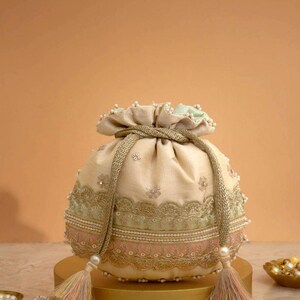 May include: A cream-colored drawstring pouch featuring intricate gold embroidery, delicate lace trim, and small white pearl accents. The bag is cinched with a thick gold cord that ends in two multi-colored tassels with square beaded embellishments.