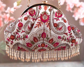 Puede incluir: Un bolso de mano blanco con una correa de cadena dorada y flecos de cuentas. El bolso está decorado con un patrón floral en tonos de rosa, rojo y verde.