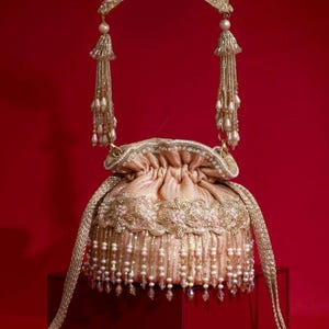 Op de afbeelding: Een perzikkleurige tas met trekkoordsluiting, voorzien van ingewikkeld goudborduursel en een brede decoratieve band. De tas is versierd met hangende parelachtige kralen en heldere kristallen, aangevuld met een bijpassend goudgeborduurd handvat.