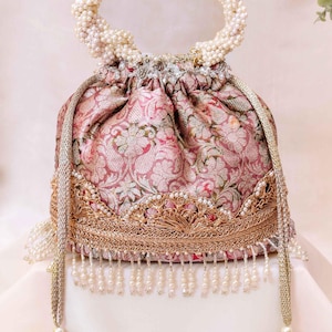 Puede incluir: Un bolso tipo saco con cordón en tela de brocado floral rosa y dorado, con un asa de perlas artificiales blancas, un borde ancho de encaje metálico dorado en la base y flecos de perlas blancas colgantes con borlas a juego en los cordones.