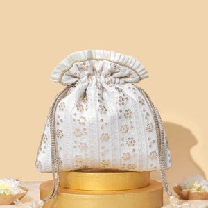May include: A white drawstring bag with gold floral embroidery and a ruffled top. The bag is sitting on two gold round platforms.