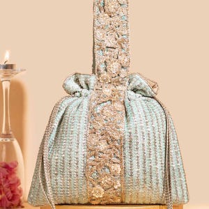 May include: A light mint green drawstring pouch bag featuring intricate gold embroidery and sequin detailing. The bag has a vertical central panel with floral patterns and a matching top handle, finished with two long braided drawstrings and tassels.