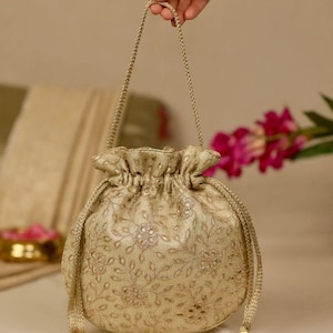 Può includere: Una borsa a sacchetto color oro con un intricato ricamo floreale in filo metallico e piccoli dettagli riflettenti. La borsa presenta una parte superiore arricciata, un manico intrecciato e due cordini laterali rifiniti con nappe di perline.
