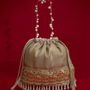 May include: A gold-colored drawstring bag with a decorative floral border and dangling pearls. The bag has a pearl-adorned handle and a red background. The bag's design includes gold embroidery and a textured fabric.