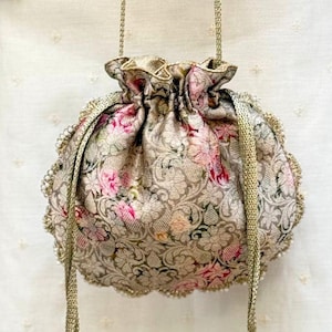 May include: A drawstring pouch bag featuring a metallic gold and beige floral brocade fabric with pink accents. The bag has a gathered top, gold braided cord handles, and decorative beaded tassels hanging from the sides with pearl-like beads.