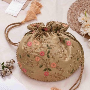 Puede incluir: Un bolso de mano dorado con cierre de cordón, decorado con intrincados bordados florales en tonos rosa y verde. El bolso presenta bordes adornados con cuentas doradas y cordones trenzados largos terminados en borlas decorativas.