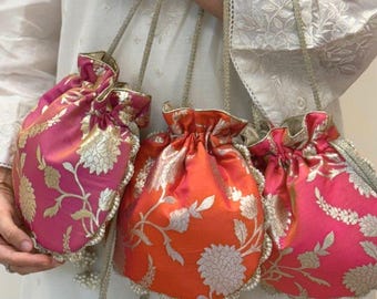 Op de afbeelding: Drie kleurrijke tassen met trekkoord en bloempatronen. De tassen zijn in roze en oranje tinten met gouden accenten. Elke tas heeft een kralenkwast en een gouden kettingriem. De tassen worden gepresenteerd tegen een witte achtergrond.
