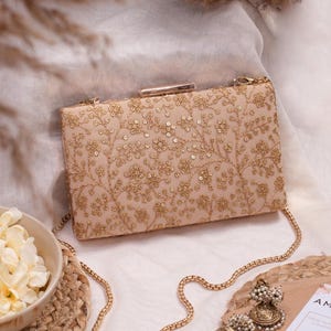 Può includere: Pochette rettangolare color cipria con ricami floreali dorati e catena dorata. La pochette è decorata con paillettes e ha una chiusura dorata. Sono visibili anche orecchini abbinati con perle.