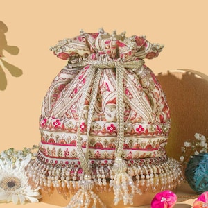 May include: A cream-coloured drawstring bag with a floral pattern in red and gold. The bag has a beaded fringe and tassel drawstrings. The background is a light peach colour.