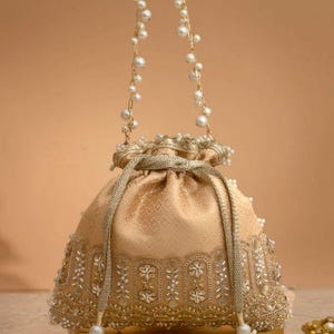 May include: A gold-toned drawstring pouch bag featuring a subtle geometric pattern, embellished with intricate beadwork and embroidery along the base. The bag includes a handle made of gold chain links accented with white pearls and matching tassels.