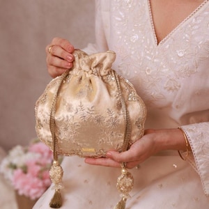 May include: A cream-coloured, drawstring pouch bag with intricate floral embroidery and gold-toned accents. The bag features a beaded tassel and a delicate chain strap. The bag is held in front of a cream-coloured outfit.