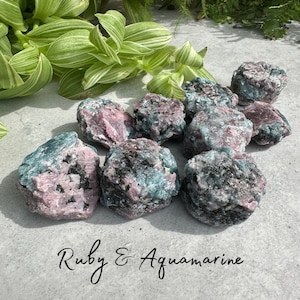 Puede incluir: Una colección de piedras rugosas de rubí y aguamarina. Las piedras muestran una mezcla de tonos rosa, azul-verdoso y negro. El texto "Ruby & Aquamarine" está escrito en cursiva en la parte inferior de la imagen.