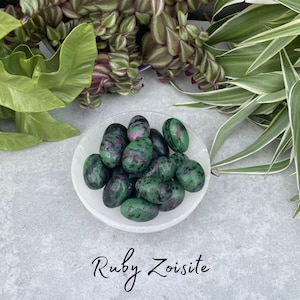 May include: A collection of green and pink ruby zoisite tumbled stones in a white bowl. The stones are smooth and polished, with a variety of shapes and sizes.  Ruby Zoisite.