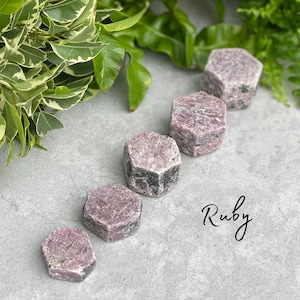 Puede incluir: Cinco cristales de rubí hexagonales de diferentes tamaños, que van desde pequeños hasta grandes, están dispuestos en una fila sobre una superficie gris. Los cristales son de color rosa intenso con inclusiones negras. La palabra "Rubí" está escrita en cursiva debajo de los cristales.