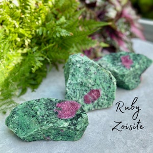 Op de afbeelding: Drie stukken ruwe groene en roze robijnzoisiet. De stenen zijn onregelmatig gevormd en hebben een gevlekt groene en roze uiterlijk. De tekst "Ruby Zoisite" is in cursief onder de stenen geschreven.