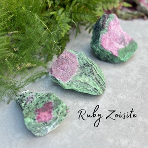 May include: Three rough, green and pink ruby zoisite stones. The stones are on a light grey surface. The text "Ruby Zoisite" is written in cursive below the stones.