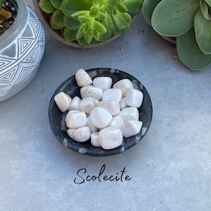 May include: A small black bowl filled with white scolecite tumbled stones. The bowl is sitting on a gray surface. Scolecite.