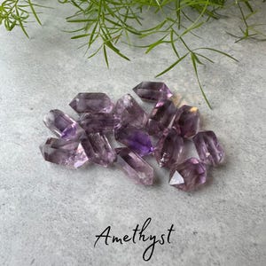 May include: A collection of amethyst crystal points, with a variety of sizes and shades of purple. The crystals are arranged on a light grey surface. Amethyst.