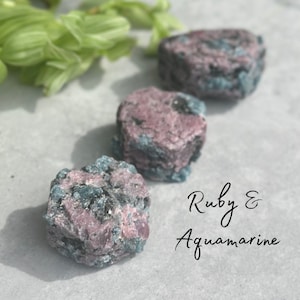 May include: Three rough, natural, pink and blue gemstones. The gemstones are labelled "Ruby & Aquamarine".