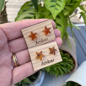 May include: Two pairs of amber star-shaped earrings displayed on wooden cards. Each card has the word "Amber" printed on it.