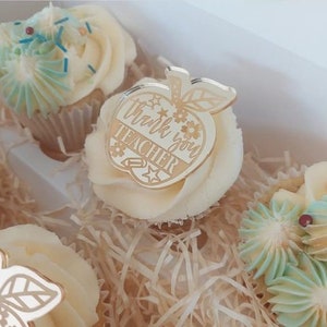 Può includere: Un set di cupcake decorati con codette blu e bianche. Un cupcake ha un topper a forma di mela in acrilico dorato con il testo "Grazie insegnante".