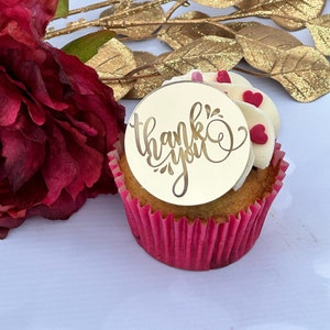 Può includere: Un cupcake bianco con glassa alla vaniglia e codette a forma di cuore rosse sormontato da un topper rotondo in acrilico dorato che dice "grazie" in corsivo.