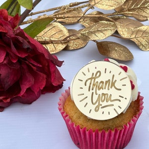 Può includere: Un cupcake con glassa bianca e un topper rotondo dorato che dice "Grazie" in corsivo. Il cupcake è in un pirottino rosa e si trova su una superficie bianca.