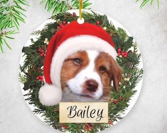 May include: A round ceramic Christmas ornament featuring a dog wearing a Santa hat. The dog is surrounded by a green holly wreath with red berries. The ornament has the name "Bailey" written on it.