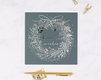 May include: A Christmas card with a wreath design. The wreath is silver and white with a bow at the top. The words "christmas wishes" are written in cursive in the center of the wreath. The card is on a gray background.