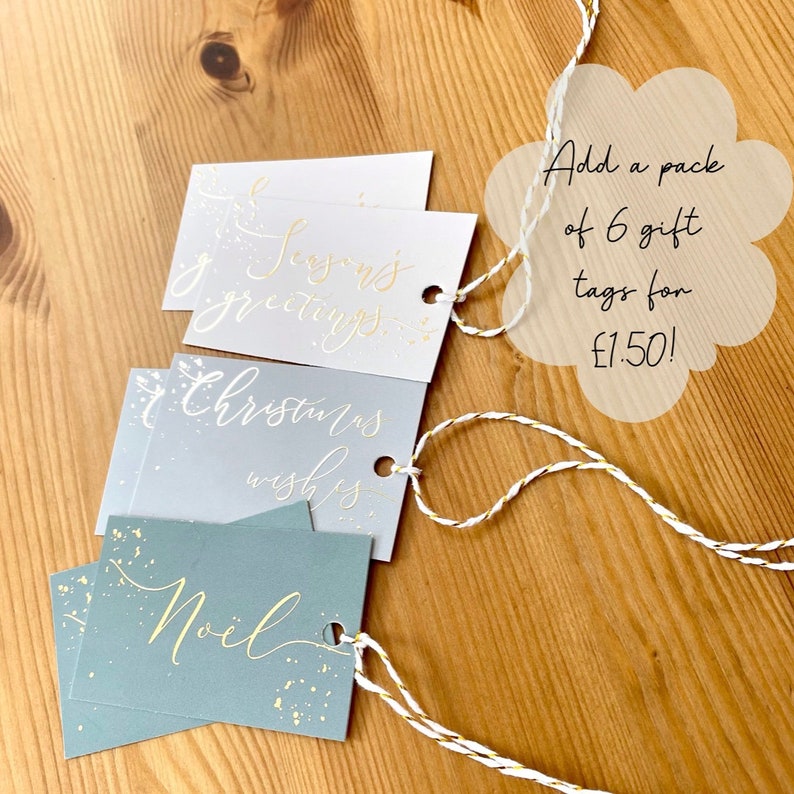 May include: Three Christmas gift tags with gold lettering on different coloured backgrounds. The tags are tied with twine and have a hole punched in the top. The text on the tags reads "Seasons Greetings", "Christmas Wishes", and "Noel".