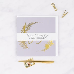 May include: A pack of six luxury Christmas cards featuring a gold foil design of a wreath on a pale purple background. The cards are packaged with a white label that reads "Paper Jewels Co. 6 Luxury Christmas Cards".