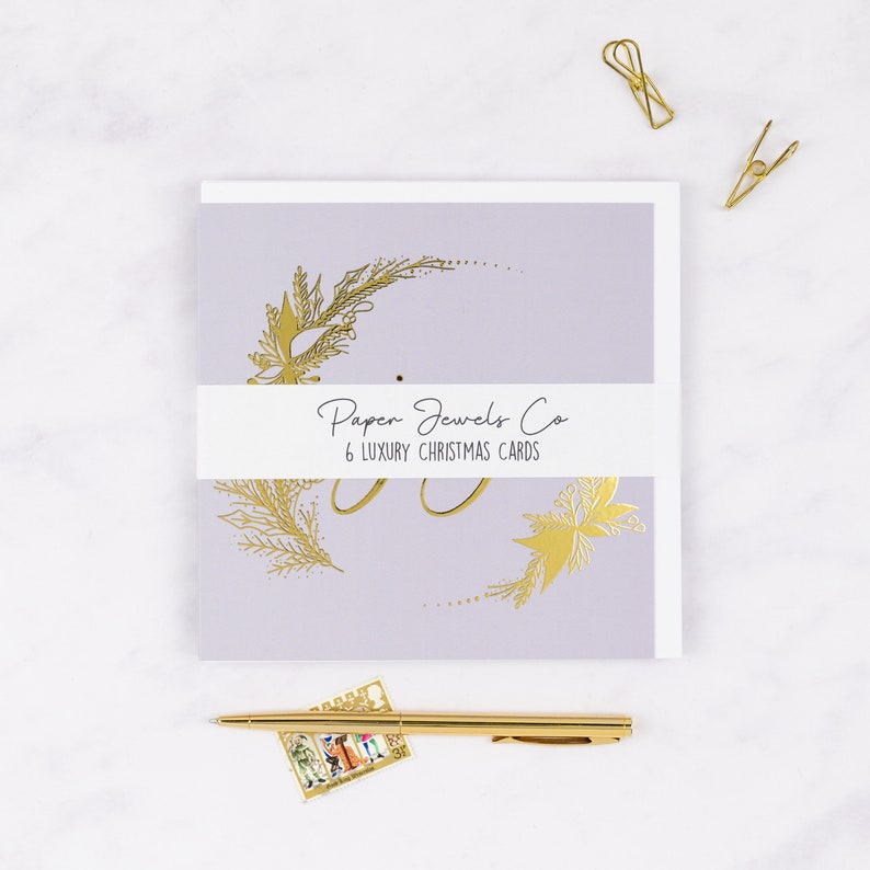 May include: A pack of six luxury Christmas cards featuring a gold foil design of a wreath on a pale purple background. The cards are packaged with a white label that reads "Paper Jewels Co. 6 Luxury Christmas Cards".