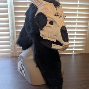 May include: A white and black skull mask with horns and black fur. The mask has a detailed skull design with dark eye sockets and a symbol on the forehead. The fur extends down the neck, creating a dramatic effect.