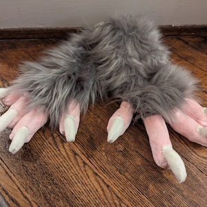 May include: A pair of plush, gray furry paws with pink pads and long, off-white claws. The faux fur is a soft, medium gray color. The claws are a light, off-white color. The paws are lying on a wooden surface.