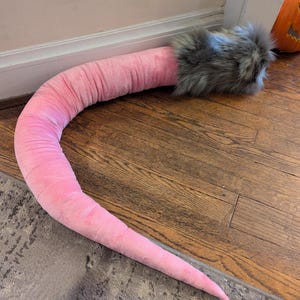 May include: A long, pink and gray plush tail with a furry tip. The tail is curled up on a wooden floor.