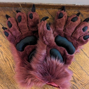 May include: A pair of burgundy-colored furry paw gloves with black paw pads and claws. The gloves are made of a soft, plush material, and the claws are pointed. The gloves are designed to resemble animal paws.