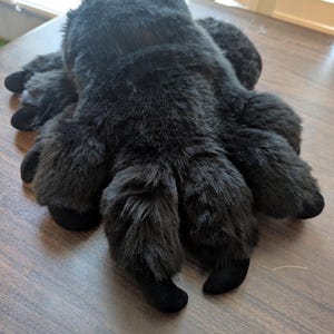 May include: A plush spider toy covered in thick, soft black faux fur. The stuffed animal features a rounded central body and eight articulated legs extending outward, each ending in a smooth, rounded black tip. The texture is dense and uniform.
