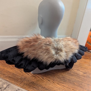 May include: A black and brown costume accessory, possibly wings, displayed on a mannequin head. The wings are black with a faux fur collar in shades of brown and tan. The item is designed to be worn and is likely for a costume or cosplay.