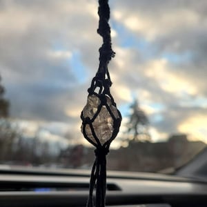 May include: A clear crystal stone is suspended in a black macrame hanging, with a cloudy sky in the background.