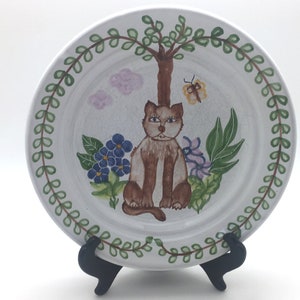 May include: A white ceramic plate with a green leafy border. The center of the plate features a hand-painted illustration of a brown cat sitting under a tree with pink clouds, blue flowers, and a yellow butterfly.