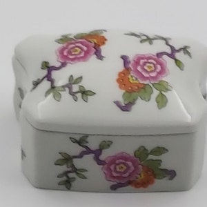 May include: A white porcelain trinket box with a floral design. The box has a square shape with rounded corners and features pink, orange, and green flowers on a white background.