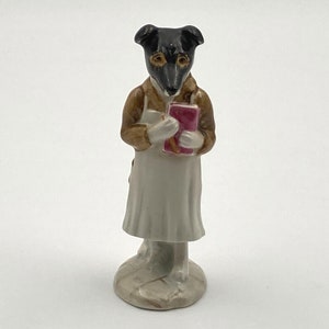 May include: A black and white dog figurine wearing a brown jacket and a white apron, holding a pink book. The figurine is standing on a white base.
