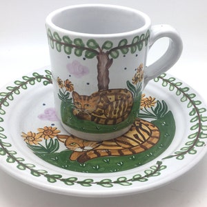 May include: A white ceramic mug and saucer set. The mug and saucer feature a hand-painted design of a brown tabby cat resting on green grass, with yellow flowers and a tree. The saucer has a green leaf border.