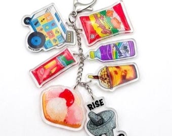 May include: A silver metal keyring featuring multiple acrylic charms including a blue food cart, a red snack bag, a purple bottle, a boba tea cup, a red candy bar, a scoop of ice cream, and a grey mortar and pestle with the text RISE AND GRIND.