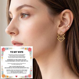 Könnte beinhalten: Goldfarbene geometrische Ohrringe mit zwei übereinanderliegenden rautenförmigen Rahmen, wobei der Name Janice in Schreibschrift auf dem unteren Rahmen eingraviert ist. Ein kleiner, klarer Ohrstecker im oberen Ohrläppchen. Eine Karte mit Text.