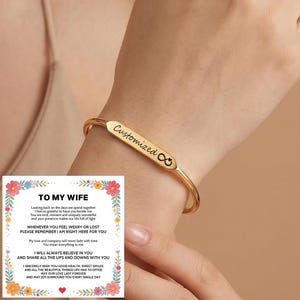 以下が含まれることがあります： Customizedという文字と無限大の記号が刻印された長方形のプレートが付いたゴールドトーンのメタルカフブレスレット。横には花柄の縁取りとTO MY WIFEという文字が印刷された白いカードが添えられています。